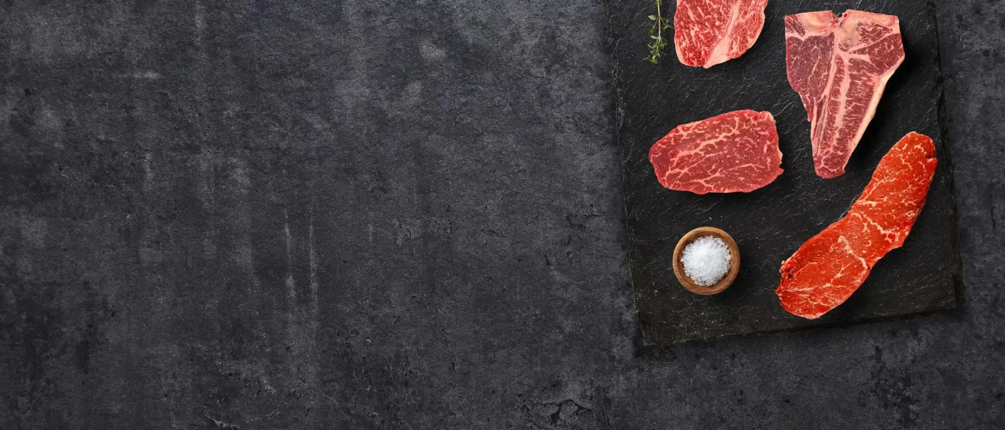Raw steaks arranged on a slate board with a small bowl of salt